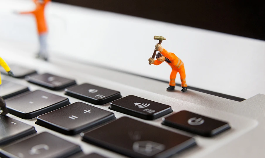 Miniature construction worker in orange overalls hammers laptop key, symbolizing tech repairs.